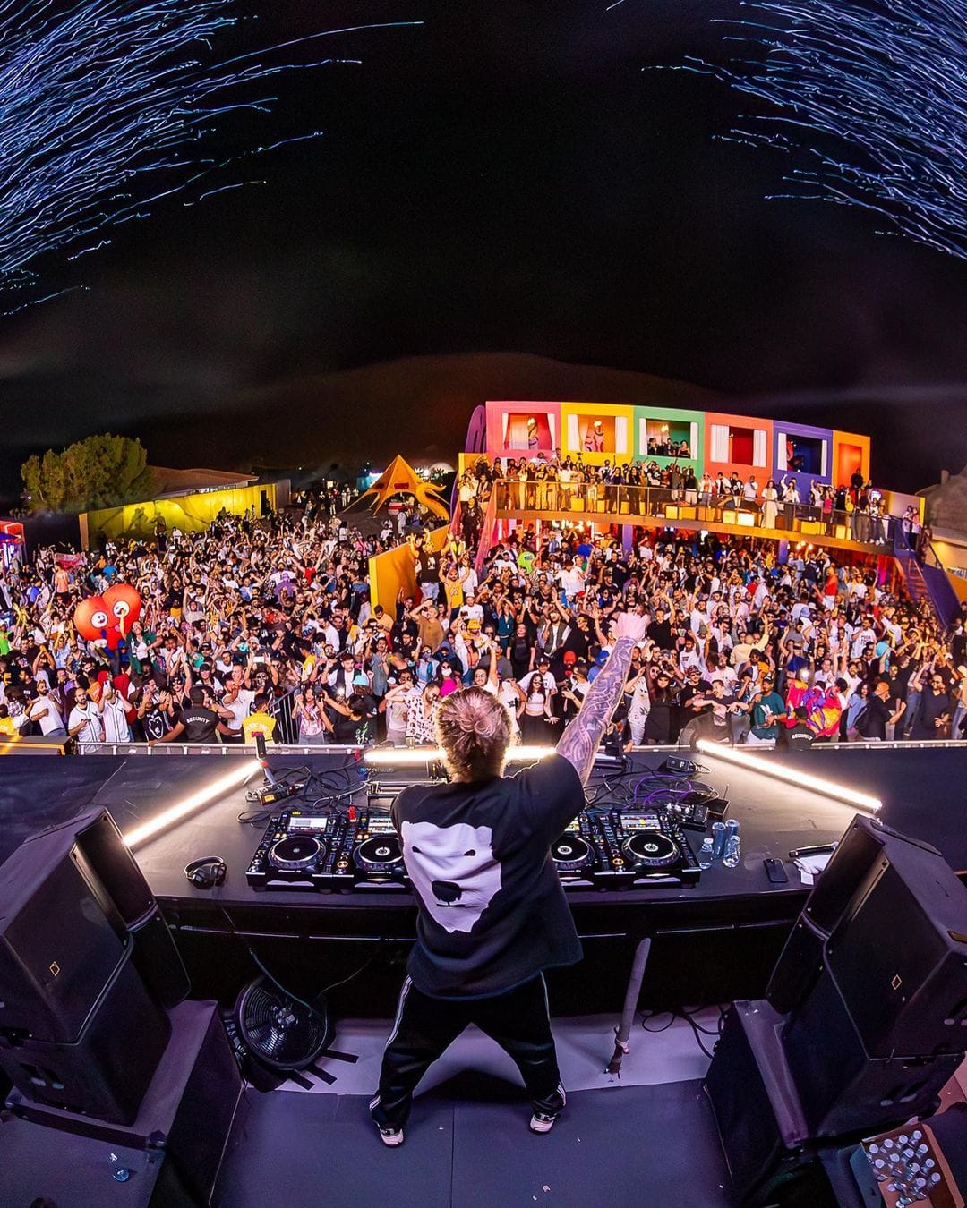 A DJ with arms raised in front of a packed festival crowd at night