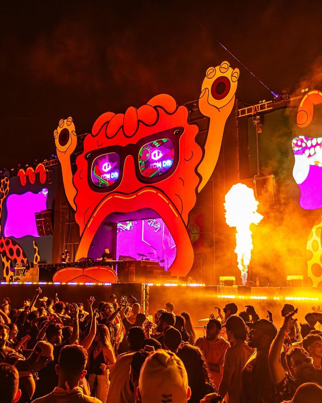 A giant cartoon-face festival stage with flame cannons firing