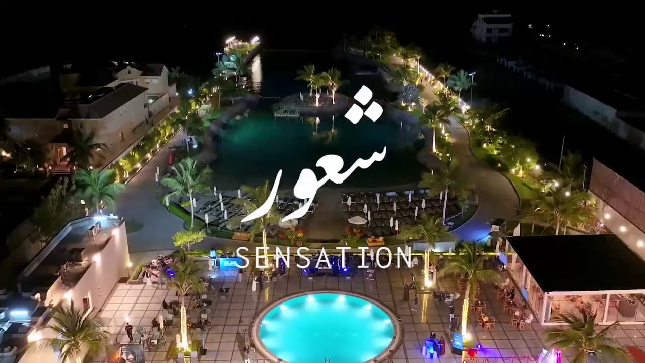 Aerial view of a beach club at night, lit pools and palms, with the Sensation logo
